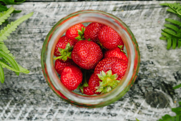 Strawberries in a glass jar on a wooden gray table on a green background, leaves of paparatnik, strawberry season.
