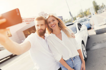 A young guy and a girl make selfie on a smartphone near their electric car.