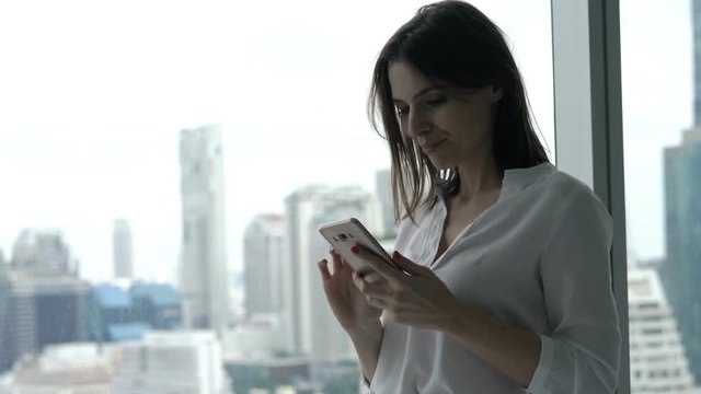 Young businesswoman texting on smartphone standing by window at office
