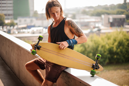 Attractive long-haired guy sitting on the parapet with longboard