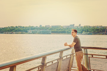 American Man summer vacation in New York. A young guy wearing green T shirt, beige pants,  standing by metal fence by Hudson River in New York opposite New Jersey. Filtered look with soft light..