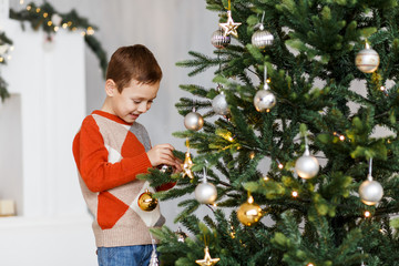 An cute boy decorating the fur-tree and fireplace with candles and gifts. New year's eve. Christmas eve. Cozy holiday at the fir-tree with lights and gold decor.