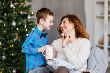 A mother and a son hugging and having fun in front of the fir-tree with candles. New year's eve. Christmas eve. Cozy holiday at the fir-tree with lights and gold decor.