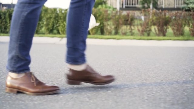 Unrecognizable man wearing blue jeans and brown leather shoes is walking in the street with no traffic. Side view. Tracking real time establishing shot