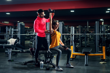 Pretty attractive fitness girl doing shoulder exercise with her personal trainer in the gym.