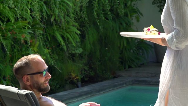Woman brings food to her boyfriend who is lying on sunbed, steadycam shot, slow motion shot at 240fps
