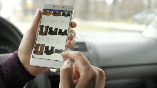 A man looks at the goods in the online shoes store, sitting in the car. Smart phone online shopping.