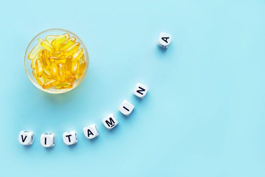 Yellow capsules in the round glass bowl and the word vitamin A from white cubes with letters on a blue background. healthy and medical concept. retinol, retinal, retinoic acid,  provitamin A