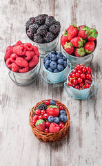Berries colorful closeup assorted mix in cans and small knitted basket. Raspberry, blueberry, red currant, strawberry on white rustic wooden table in studio.