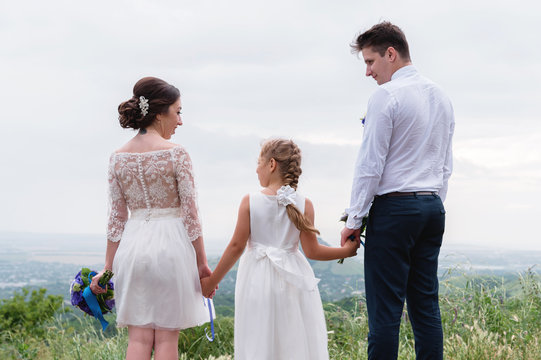 young married couple and their little daughter on wedding day are standing in nature holding hands