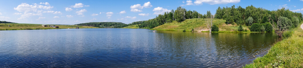 Fototapeta na wymiar The hilly coast of the lake with trees and meadows, a place to relax on a summer day. panorama