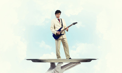 Naklejka premium Businessman on metal tray playing electric guitar against blue sky background