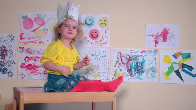 Angry beautiful kid eating cookie sitting on table against paintings on wall