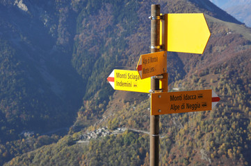 Wegweiser aus gelb beschichtem Aluminium an Wanderweg an den Berghängen östlich des Lago Maggiore, Tessin, Italien