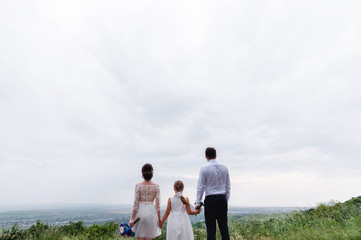 young married couple and their little daughter on wedding day are standing in nature holding hands