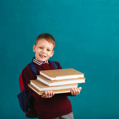 Cheerful thoughtful little school boy in school uniform with backpack and big pile of books standing against blue wall. School concept. Back to School