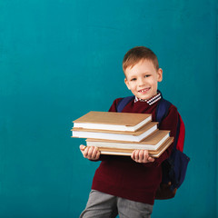 Cheerful thoughtful little school boy in school uniform with backpack and big pile of books standing against blue wall. Looking at camera. School concept.
