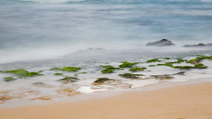 Beach rocks covered with green moss with silky waves from long exposure