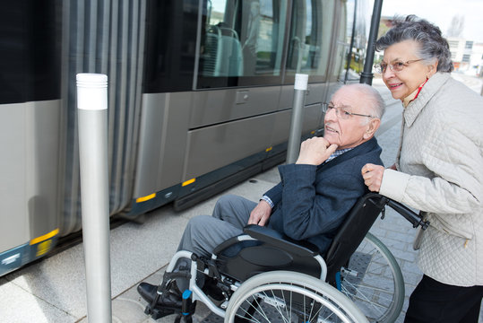Elderly woman pushing wheelchair bound husband onto tram