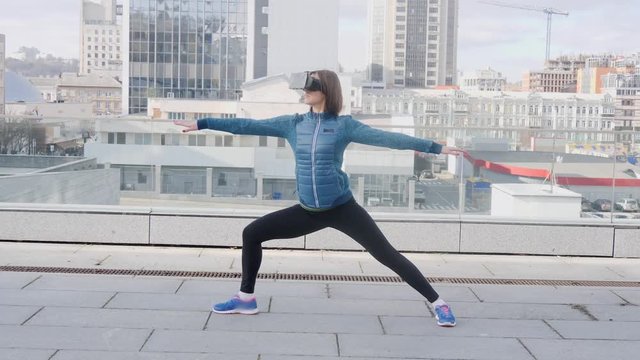 Woman practicing yoga fitness exercise at city background with office buildings wearing vr virtual reality headset.