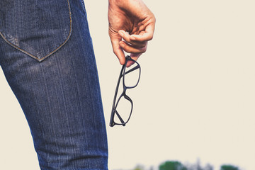 a person wearing blue jeans, holding a pair of black eyeglasses in their right hand.