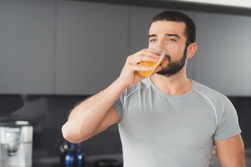 Fototapeta premium A sporty man stands in the kitchen and drinks orange juice. Next to him on a stack of vegetables and fruits.