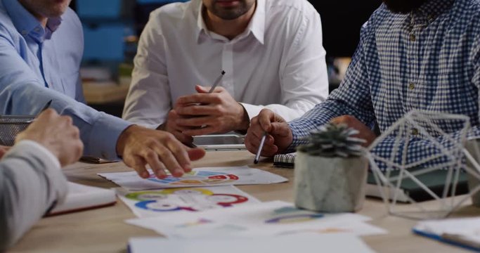 Attractive male office workers sitting at the table and discussing seriously their work with a boss in the comfortable office late in evening. Indoors. Close up of table with documents and charts