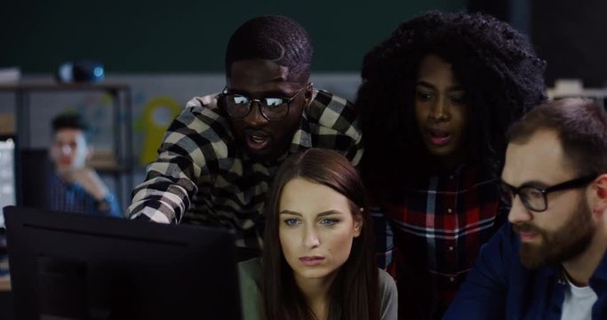 Young multi-ethnical team of office workers having a discussion about a startup project late at night. Group of friendly proffesionals looking at the computer in the urban office space.