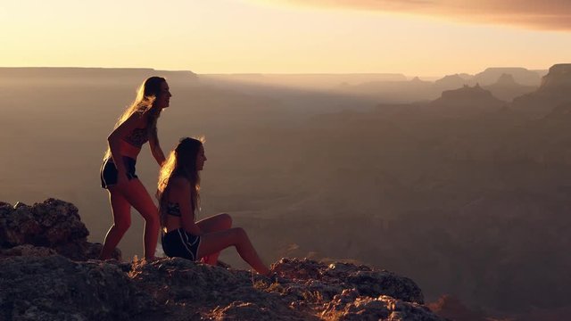 Two young women enjoying sunset view as one lends had to other to help her stand up and safely walk away from the egde of the Grand Canyon.
