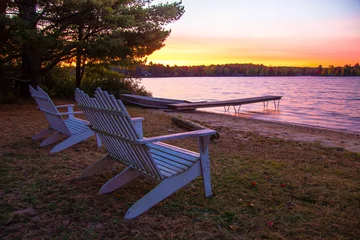 Papier Peint photo Lac / étang Vacances d& 39 été au bord du lac. Coucher de soleil sur un lac avec une rangée de chaises Adirondack, un quai et une barque en aluminium.