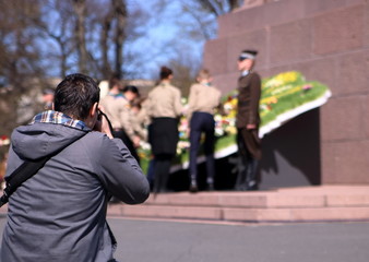 Fotograf, tyłem, fotografuje miejsce pamięci w Rydze, Łotwa, podczas uroczystości święta narodowego, w tle niewyraźny żołnierz pełniący wartę przy mapie Łotwy zrobionej z kwiatów, młodzież