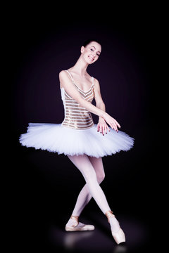 Young female / woman / girl ballerina in a white pack / tutu solo dancing and graceful staying in dark black scene with reflecting floor and dark background