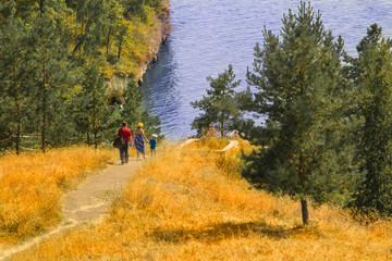 Obraz premium A man with a bag, a woman in a dress, a hat and a bag and a child in shorts, a T-shirt and a cap walking along a sand trail among the yellow grass and green trees to the river bank.