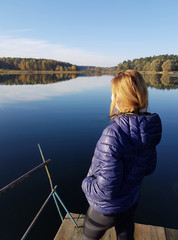 a girl in a blue jacket is standing alone on a wooden pier by the lake on an autumn day