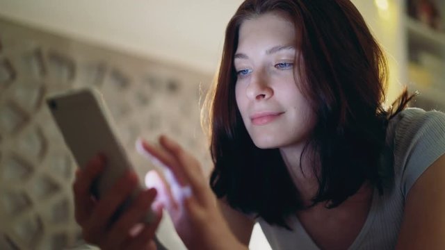 Close-up of young smiling woman using smartphone lying in bed at home at night