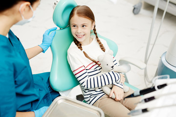 Little girl with teddybear sitting on armchair in dentist clinics and looking at doctor