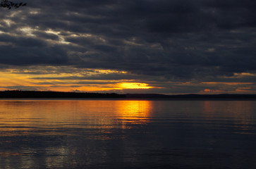 Fototapeta premium Sunset sky panorama. Bright sun line against dark clouds background. Reflection on a water.