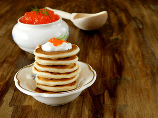 Red caviar in a white ceramic bowl and pancakes with caviar on a wooden table