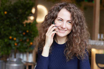 Indoor shot of stylish attractive woman with curly hair in blue sweater, calls her friend over smart phone, sits in restaurant, smiling charmlike, looks at window. Technology and communication.