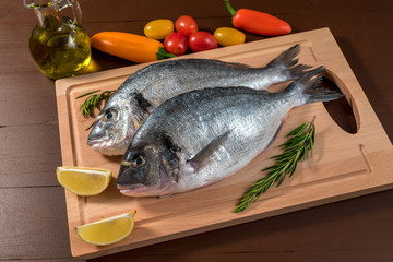 Fresh uncooked dorado or sea bream fish with lemon, herbs, oil, vegetables and spices on wooden board over dark backdrop, top view.