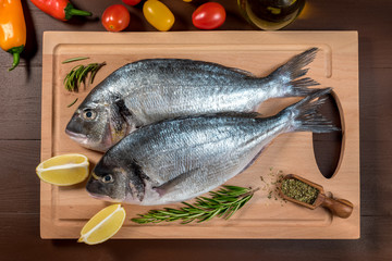 Fresh uncooked dorado or sea bream fish with lemon, herbs, oil, vegetables and spices on wooden board over dark backdrop, top view.