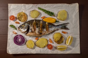 Tasty baked whole fish on baking paper. Baked sea bream with lemon, onion, herbs, cherry tomatoes, spices on dark rustic background. Grilled delicious fish. Diet and healthy food. Top view