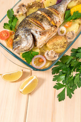 A delicious whole baked fish. Baked sea bream with lemon, onions, herbs, cherry tomatoes, spices and baked potatoes on a wooden background. Fried tasty fish. Diet and healthy food. Top view.