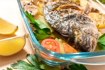 A delicious whole baked fish. Baked sea bream with lemon, onions, herbs, cherry tomatoes, spices and baked potatoes on a wooden background. Fried tasty fish. Diet and healthy food.