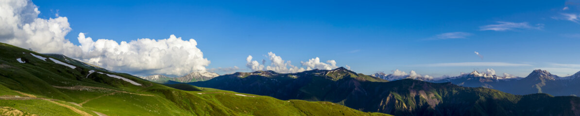 Obraz premium Wide mountain panorama.Green hills with snow on peaks in evening sunlight. Dark shadows. Blue sky with some clouds. Summer in Georgia.