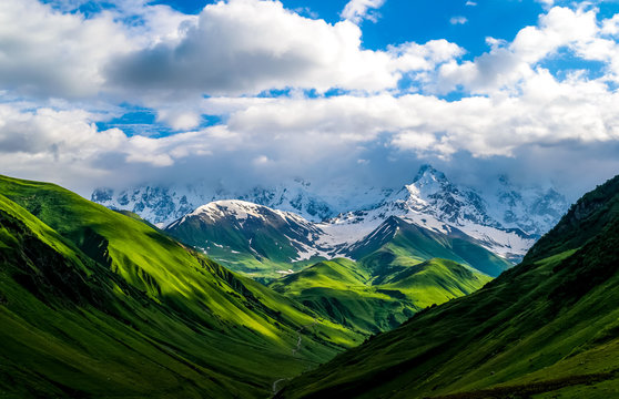 Beautiful landscape of mountains and valleys. Green fields and hills leading to snow-covered mountains. White clouds on the mountain range. Summer in Caucasian. Georgia.