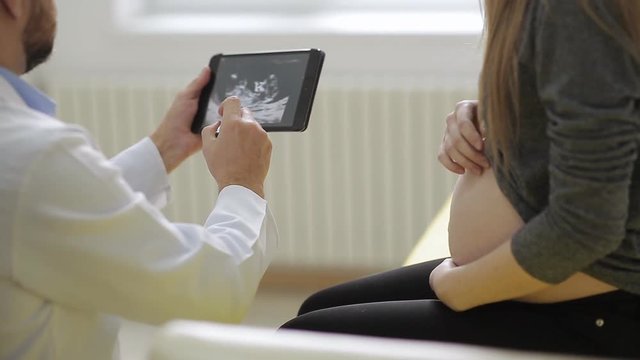 male doctor in medical robe shows a pregnant woman a picture of an ultrasound on a tablet