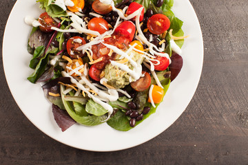 
Green spinach salad with chopped red and orange tomato, black beans, shredded cheese, avocado, and salsa on a white plate above shot
