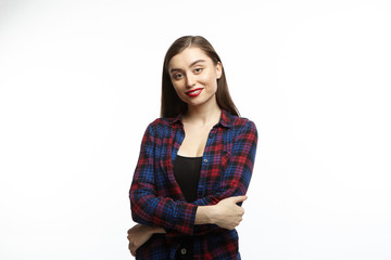Beautiful positive girl with long dark hair, wearing red lipstick and plaid shirt smiling happily and posing with arms folded against white studio wall background with copy space for your information