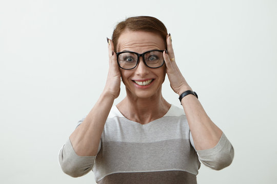 Indoor portrait of middle age woman in glasses with smile on the happy face. Female teatcher look into the camera with wide open eyes and keep head by hands. Laughing housewife